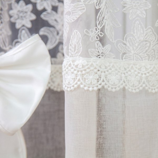 Zbliżenie na górną część firany Estella, prezentujące szeroki pas haftu o motywie kwiatowym oraz wykończenie krawędzi koronką.